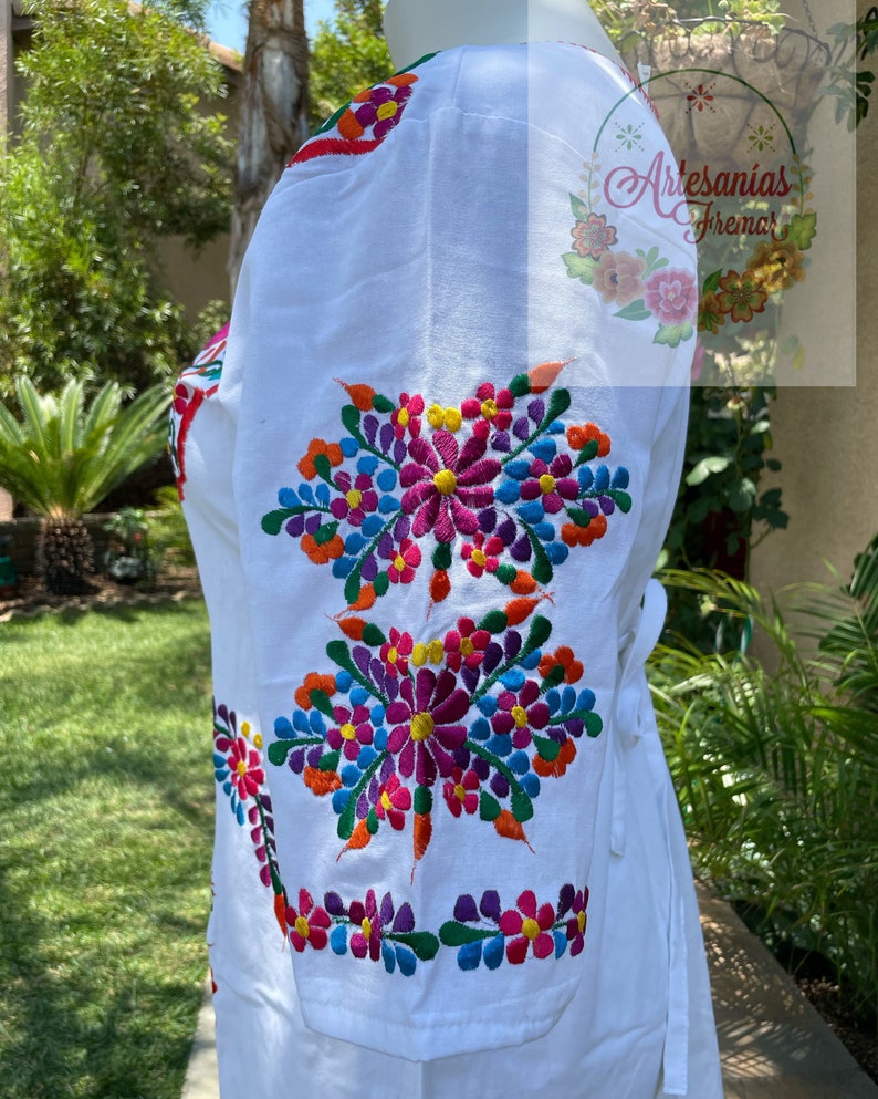 May include: White embroidered blouse with colorful floral designs. The blouse has a tie at the waist and three-quarter sleeves.