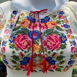 May include: White embroidered blouse with a floral pattern in shades of pink, blue, yellow, and green. The blouse has a gathered neckline and short sleeves.