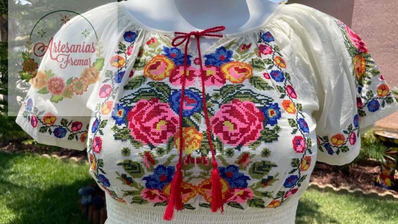 May include: White embroidered blouse with a floral pattern in shades of pink, blue, yellow, and green. The blouse has a gathered neckline and short sleeves.