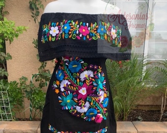 May include: Black off-the-shoulder dress with vibrant floral embroidery. The dress features colorful flowers in shades of pink, blue, white, and green. The embroidery is detailed and covers the neckline, bodice, and hemline. The dress is displayed on a mannequin.