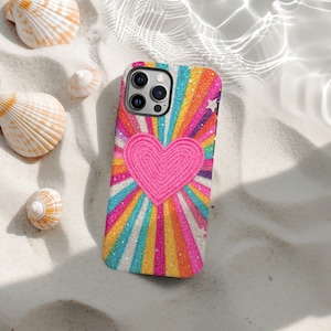 May include: A phone case with a bright design, featuring a pink heart at the centre, with rainbow stripes radiating outwards. The case has a glittery finish and small white stars. Seashells are scattered around the phone case on a sandy surface.