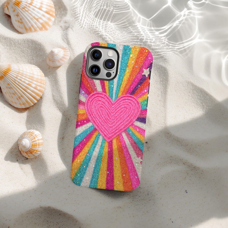 May include: A phone case with a bright design, featuring a pink heart at the centre, with rainbow stripes radiating outwards. The case has a glittery finish and small white stars. Seashells are scattered around the phone case on a sandy surface.