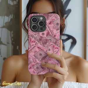 May include: A pink phone case featuring a repeating pattern of pink $100 bills and glitter accents. The case has a camera cutout and the text "Dreams Case" in the bottom left corner.