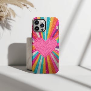 May include: A phone case with a bright, eye-catching design. It features a pink heart at the centre, set against a backdrop of rainbow-coloured rays. The case has a glittery texture, adding a touch of sparkle. Designed for a smartphone.