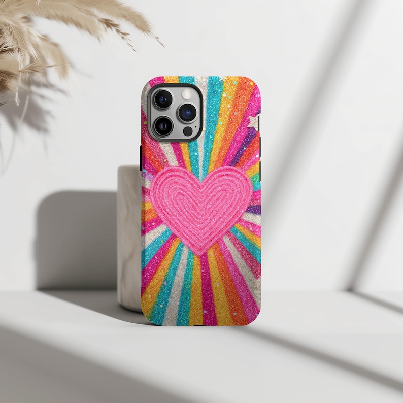 May include: A phone case with a bright, eye-catching design. It features a pink heart at the centre, set against a backdrop of rainbow-coloured rays. The case has a glittery texture, adding a touch of sparkle. Designed for a smartphone.