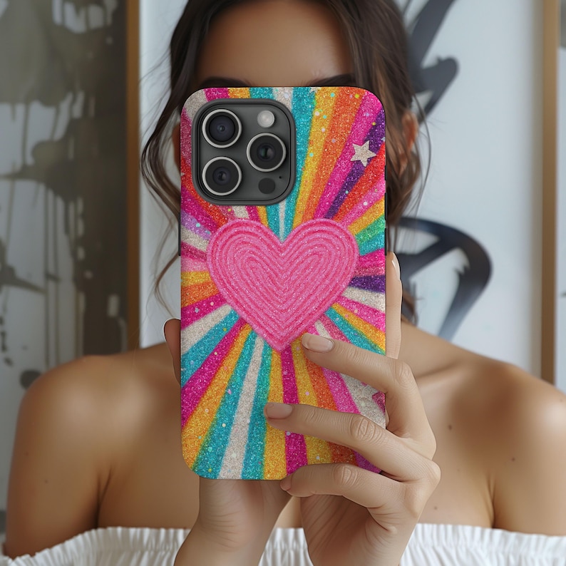 May include: A brightly coloured phone case with a heart design. The case has a pink heart at the centre, with rainbow-coloured rays radiating outwards. A small white star is visible. The case is designed for a smartphone with multiple camera lenses.