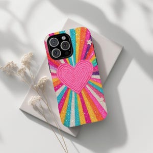 May include: A colourful phone case with a pink heart design. The case features a vibrant rainbow burst pattern with glitter accents and small white stars. The phone case is designed to fit a smartphone.