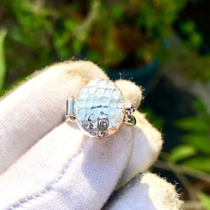 May include: A silver ring with an oval, textured, light blue gemstone. The ring has a hammered metal design and is held by a gloved hand. The background is blurred with green foliage.