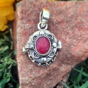 May include: A silver locket pendant featuring a red gemstone. The locket has an intricate design and a hinged opening.