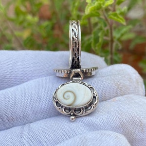 May include: A silver ring with a white shell pendant. The ring has a braided design and the pendant is oval shaped with a swirl pattern.