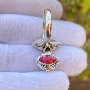 May include: A silver ring with a red gemstone. The ring features a decorative setting around the gemstone and a detailed band. The gemstone is an oval shape and is a vibrant red color. The ring is held against a white background.