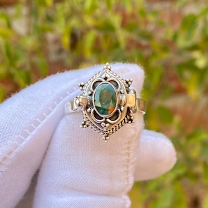 May include: A silver ring with an oval, green gemstone at the center. The ring has an ornate, filigree design around the gemstone and on the band. The ring is held up by a gloved hand.