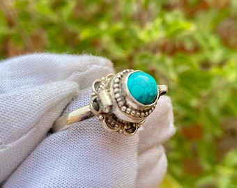 Könnte beinhalten: Ein silberner Ring mit einem türkisfarbenen Stein, der in einer kleinen, verzierten Schachtel sitzt. Der Ring liegt auf einer weißen Oberfläche.