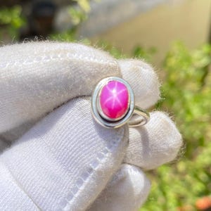 Może przedstawiać: Srebrny pierścionek z różowym kamieniem szlachetnym rubinu gwiazdowego. Owalny rubin jest osadzony w oprawie. Pierścionek jest trzymany w białej rękawicy na tle zielonych liści. Widoczny jest efekt gwiazdy rubinu.