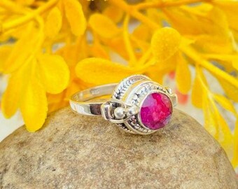 Könnte beinhalten: Ein silberner Ring mit einem rosafarbenen Edelstein, der in einer dekorativen Fassung sitzt. Der Ring liegt auf einem grauen Stein mit gelben Blumen im Hintergrund.