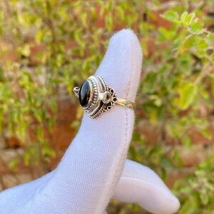May include: A silver ring with an oval, black gemstone. The ring has an ornate design around the stone and on the band, with a detailed, textured pattern. The ring is displayed on a white surface.