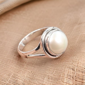 May include: A silver ring with a white pearl set in a circular bezel. The ring has a split band design.
