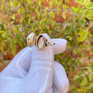 May include: Open silver locket ring with an oval-shaped compartment. The ring features a decorative design around the compartment and a small clasp. The locket is held open, revealing the interior.