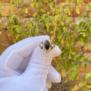 May include: A silver ring with an oval, black gemstone. The ring has an ornate design around the gemstone and on the band. The ring is held up by a gloved hand, with a blurred background of green leaves and a brick wall.