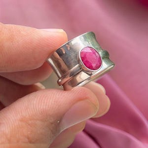 May include: A silver ring with a large, oval-shaped, pink gemstone set in the center. The ring is wide and has a textured, hammered finish.