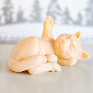 Puede incluir: Una pequeña figurita de cerámica de color crema que representa a un gato durmiendo con una pata levantada en el aire.