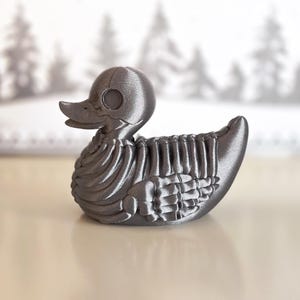Puede incluir: Un pato de goma impreso en 3D con un diseño esquelético. El pato está hecho de plástico gris oscuro y tiene una estructura esquelética detallada.