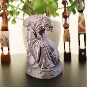 Pode incluir: Uma figurinha de Buda sorridente com cabeça de alienígena impressa em 3D na cor roxa. A figurinha está sentada em uma base com um design em espiral.
