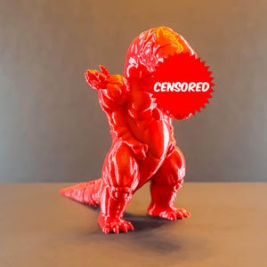 Könnte beinhalten: Eine rote 3D-gedruckte Figur eines Monsters mit einem zensierten Gesicht. Das Monster steht auf einer grauen Oberfläche.