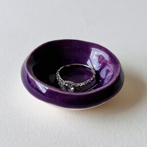 Puede incluir: Un pequeño cuenco redondo de cerámica con un esmalte morado oscuro. Un anillo de plata con una banda decorativa y una piedra central descansa dentro del cuenco. El cuenco tiene un acabado liso y brillante y una parte inferior blanca.