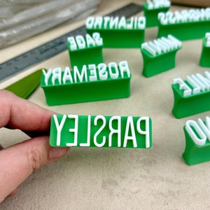 May include: A collection of green and white plastic herb stamps featuring raised lettering for labels including Parsley, Rosemary, Sage, Cilantro, Lemongrass, Mint, Thyme, Chamomile, and Oregano, designed for imprinting into clay or soft materials.