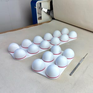 Puede incluir: Dos bandejas rectangulares blancas con múltiples cúpulas hemisféricas blancas, cada una acentuada con un fino anillo rojo en la base. Una herramienta de metal delgada de color plateado con punta está colocada junto a la bandeja pequeña.