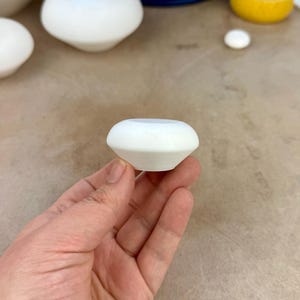 Puede incluir: Un pequeño recipiente de cerámica en color blanco mate con un cuerpo redondeado y cónico, y una parte superior abierta y ancha. El objeto presenta un acabado liso y uniforme con una forma geométrica que se estrecha hacia la base.