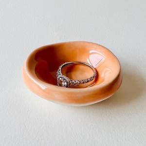 Könnte beinhalten: Eine kleine, pfirsichfarbene Keramik-Ring Schale mit weißem Innenraum. Ein silberner Ring mit einem großen, klaren Edelstein liegt in der Schale.