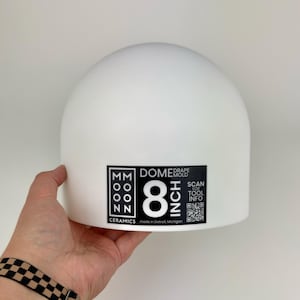 May include: A white ceramic dome drape mold with a black label featuring the text Moon Ceramics, 8 Inch Dome Drape Mold, and a QR code. The mold has a smooth, rounded top surface and a cylindrical base, designed for pottery and clay forming projects.
