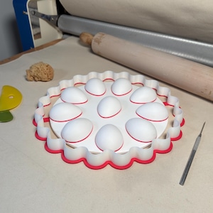 May include: A circular white and red plastic mold featuring a scalloped edge and nine raised, egg-shaped domes arranged in a circular pattern. The mold is designed for shaping materials, with a red base rim and a white top surface.