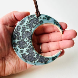 May include: A ceramic crescent moon ornament with a teal glaze and black speckles. The ornament has a brown leather cord for hanging.