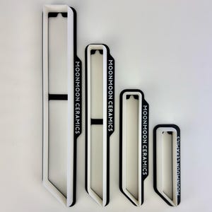 Puede incluir: Cuatro piezas de cerámica blancas y negras de diferentes tamaños. Cada pieza tiene forma rectangular con la parte superior e inferior curvadas. El texto "MOONMOON CERAMICS" está impreso verticalmente en el lateral de cada pieza.