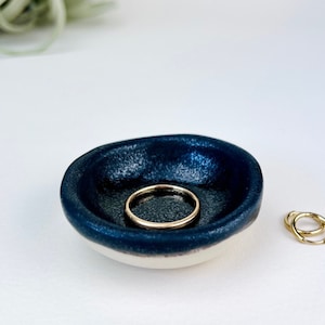 May include: A small, dark blue ceramic ring dish with a white rim. The dish is slightly irregular in shape and has a textured surface. Two gold rings are sitting on the dish.