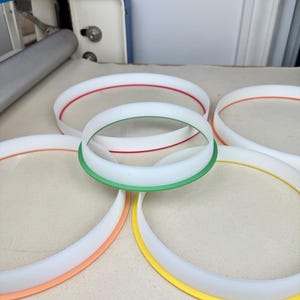 May include: Five white plastic embroidery hoops with colored trim. The hoops are arranged in a circle, with the largest hoop at the top and the smallest hoop at the bottom. The hoops are red, green, orange, and yellow.