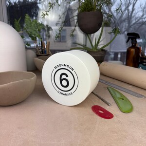 May include: A cylindrical white ceramic tool with a flat face featuring a black circular logo. The logo contains the text MOONMOON CERAMICS surrounding the number 6. The object is positioned on a flat surface alongside various pottery tools.
