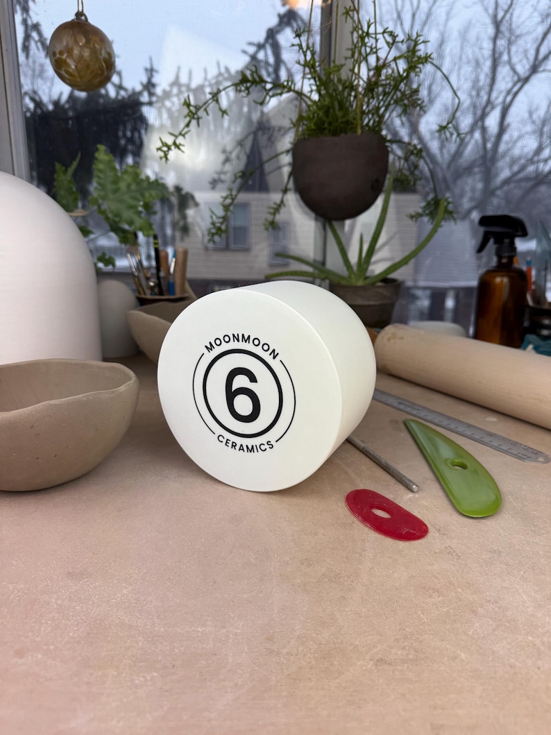May include: A cylindrical white ceramic tool with a flat face featuring a black circular logo. The logo contains the text MOONMOON CERAMICS surrounding the number 6. The object is positioned on a flat surface alongside various pottery tools.