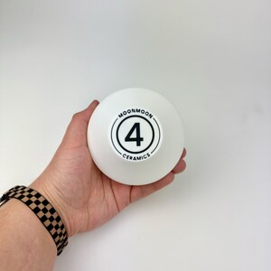 May include: A white ceramic bowl with a smooth, matte finish. The center of the bowl features a black circular logo containing the number 4, surrounded by the text MOONMOON CERAMICS in a clean, sans-serif font, all printed in black ink.