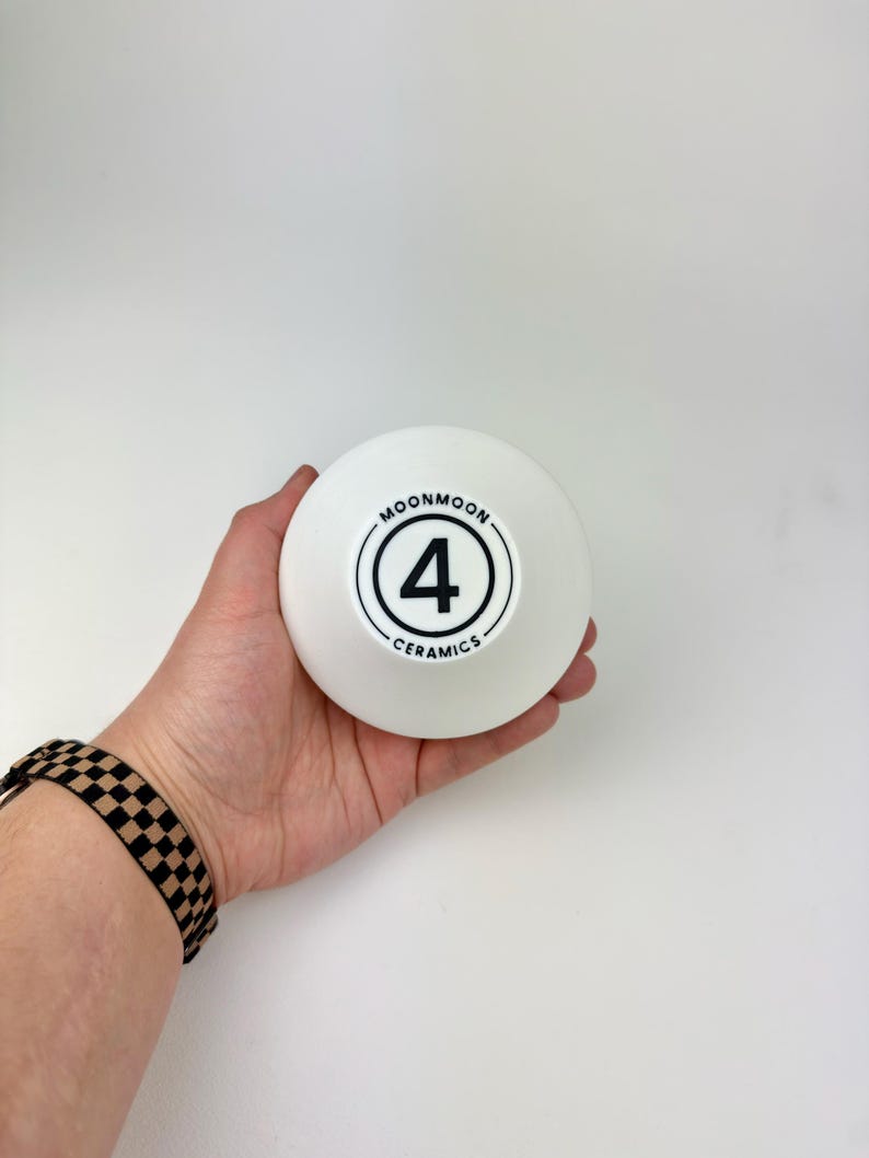 May include: A white ceramic bowl with a smooth, matte finish. The center of the bowl features a black circular logo containing the number 4, surrounded by the text MOONMOON CERAMICS in a clean, sans-serif font, all printed in black ink.