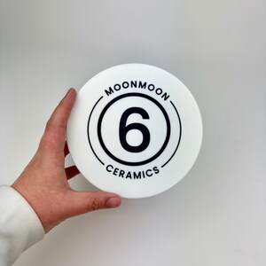 May include: A circular white ceramic object featuring a black graphic design. The center displays the number 6 enclosed within a circular border. The text MOONMOON is printed along the top curve and CERAMICS is printed along the bottom curve.