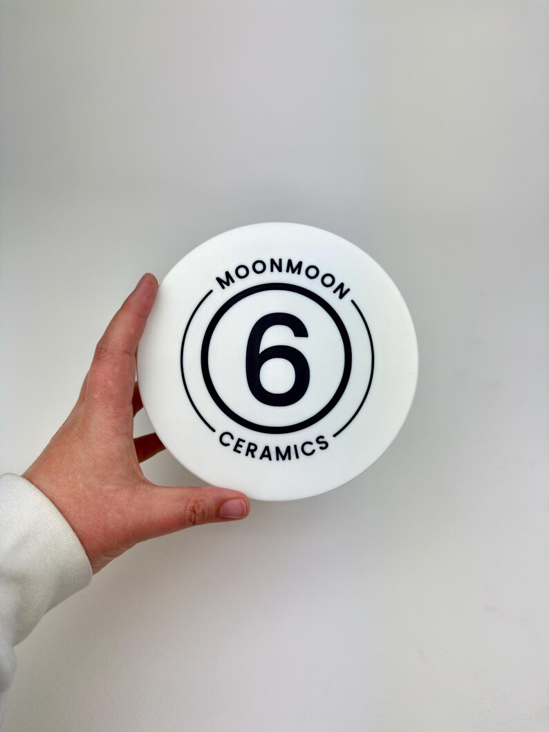 May include: A circular white ceramic object featuring a black graphic design. The center displays the number 6 enclosed within a circular border. The text MOONMOON is printed along the top curve and CERAMICS is printed along the bottom curve.