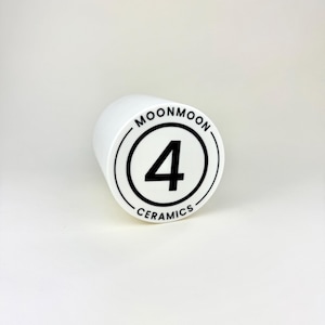 Op de afbeelding: Een witte keramische cilinder met de tekst "MOONMOON" en "CERAMICS" in een zwarte cirkel, met het nummer "4" in het midden. De cilinder staat op een witte achtergrond.