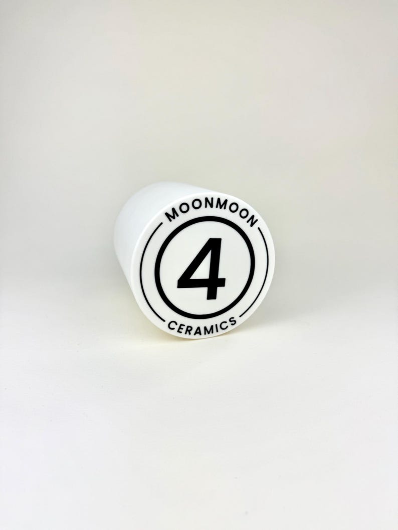 Op de afbeelding: Een witte keramische cilinder met de tekst "MOONMOON" en "CERAMICS" in een zwarte cirkel, met het nummer "4" in het midden. De cilinder staat op een witte achtergrond.