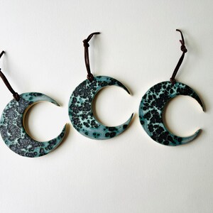 May include: Three ceramic crescent moon ornaments with a teal and black speckled glaze. Each ornament is attached to a brown leather cord.