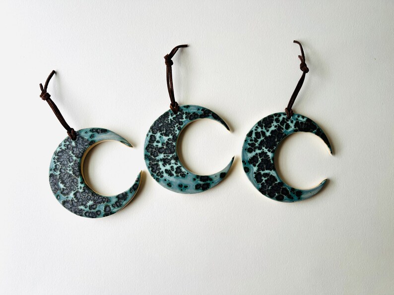 May include: Three ceramic crescent moon ornaments with a teal and black speckled glaze. Each ornament is attached to a brown leather cord.