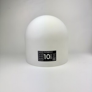 May include: A white dome drape mold with a smooth, matte surface. A black rectangular label on the front displays the text Moon Ceramics, Dome Drape Mold, 10 Inch, and includes a QR code for tool information. The object has a rounded top.